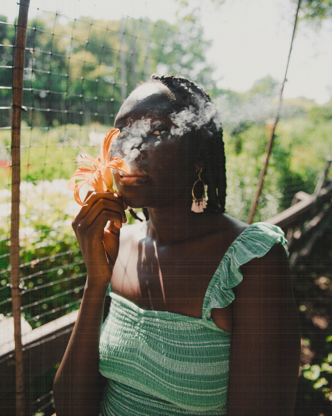Photo of a Black woman in a green top exhaling smoke from and holding an orange flower near her mouth.